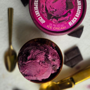 Graeter's Black Raspberry Chip ice cream container with a scoop removed, surrounded by chocolate pieces and a spoon.