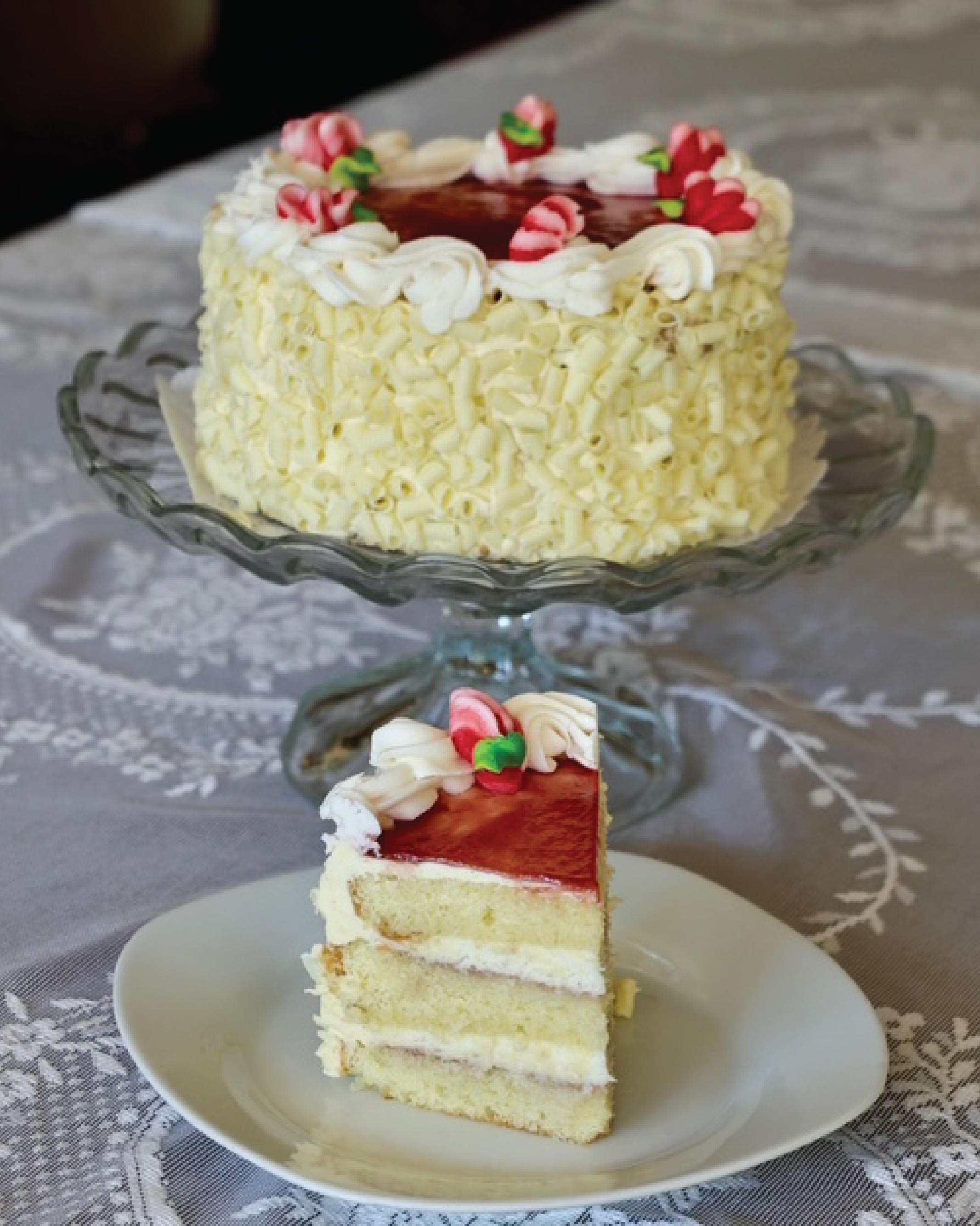 A white chocolate bumbleberry cake with red fruit topping is displayed on a glass pedestal stand, with a single slice placed on a white plate in front. The cake is decorated with piped white frosting and red-and-green candy accents, set on a patterned gray tablecloth.