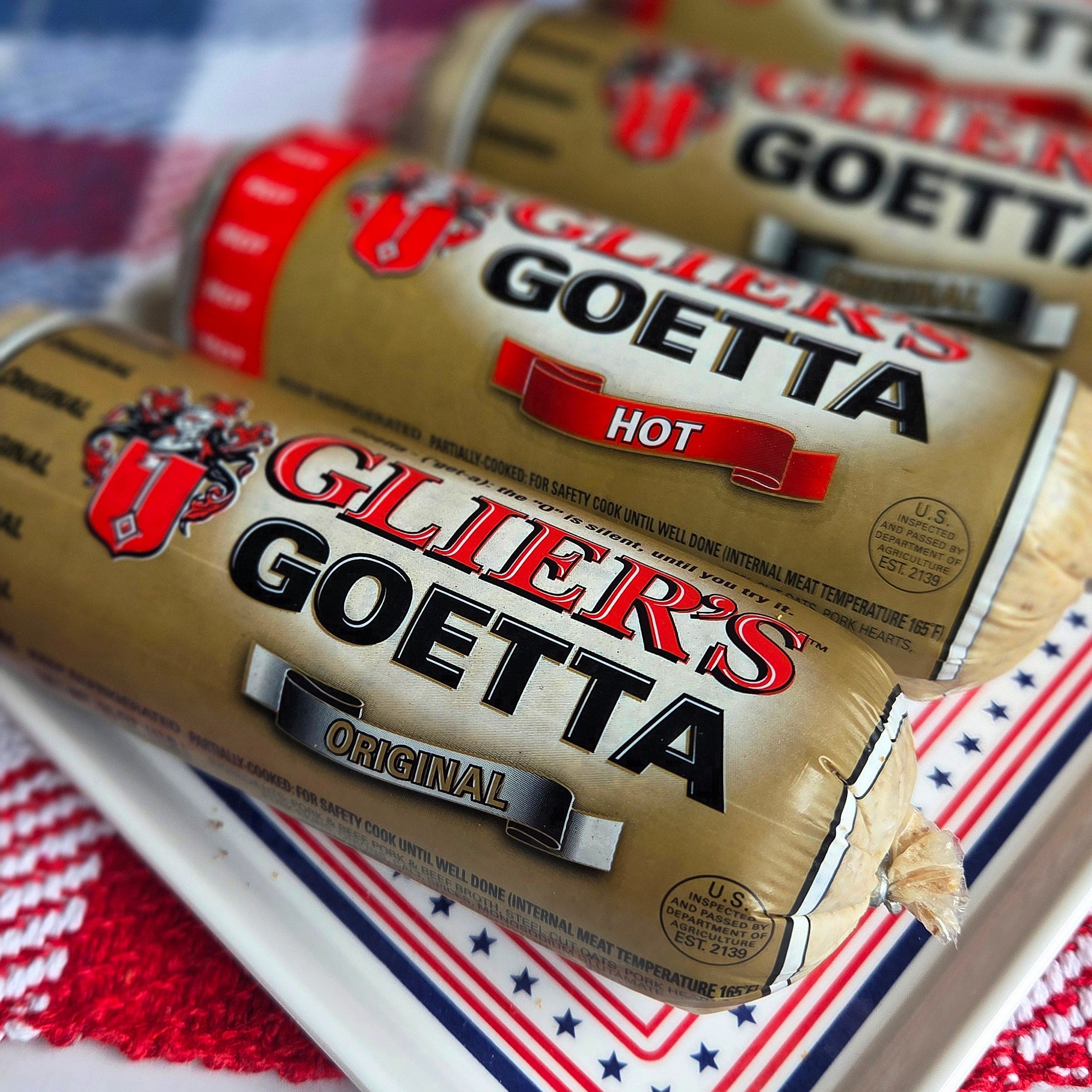 Three packages of Glier's Goetta Original hot sausage rolls arranged on a white serving tray with a red, white, and blue checkered cloth underneath.