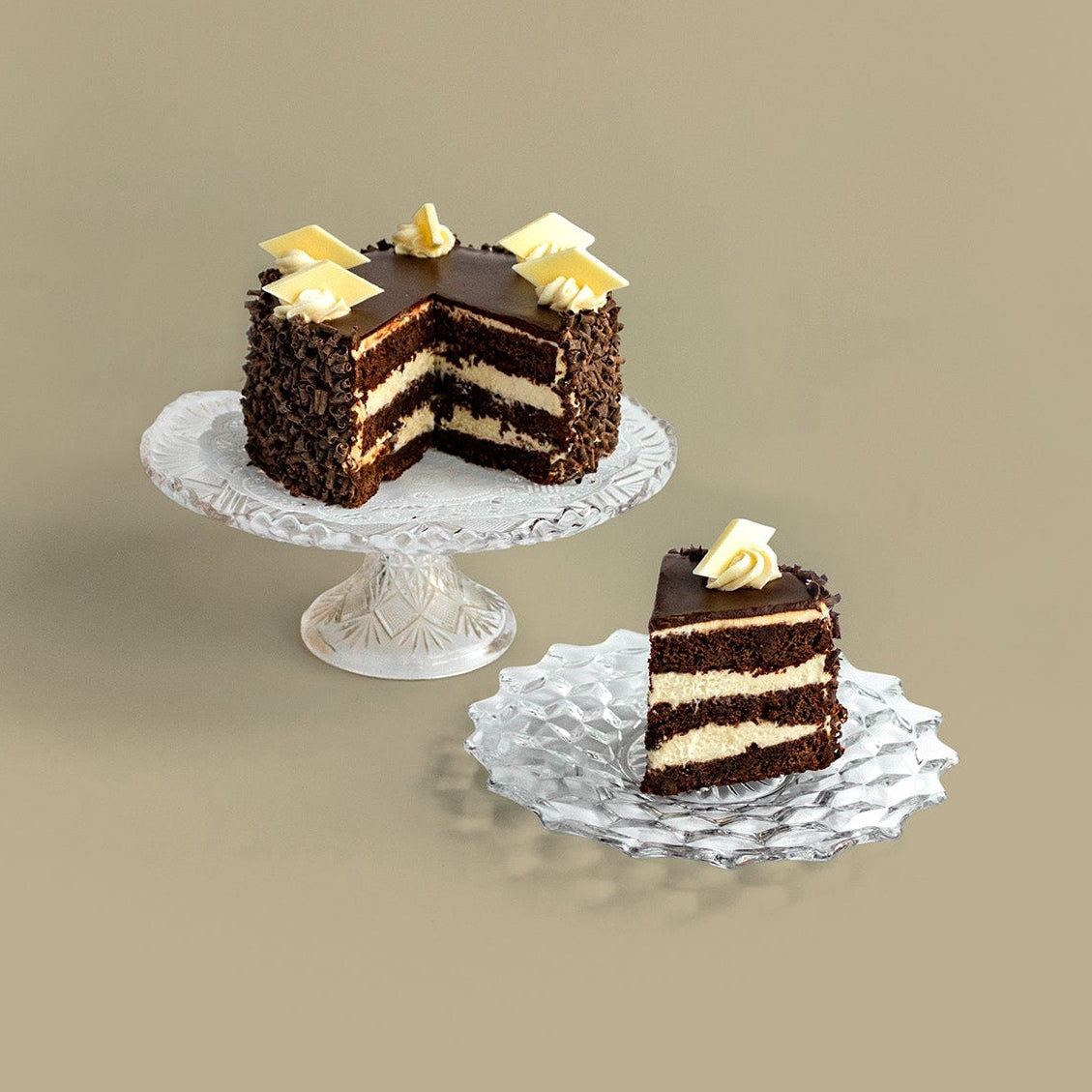 A BONBONERIE OPERA CREAM cake with cream-colored layers and yellow flower decorations sits on a white pedestal, with a single slice placed on a textured silver plate in front, set against a solid beige background.