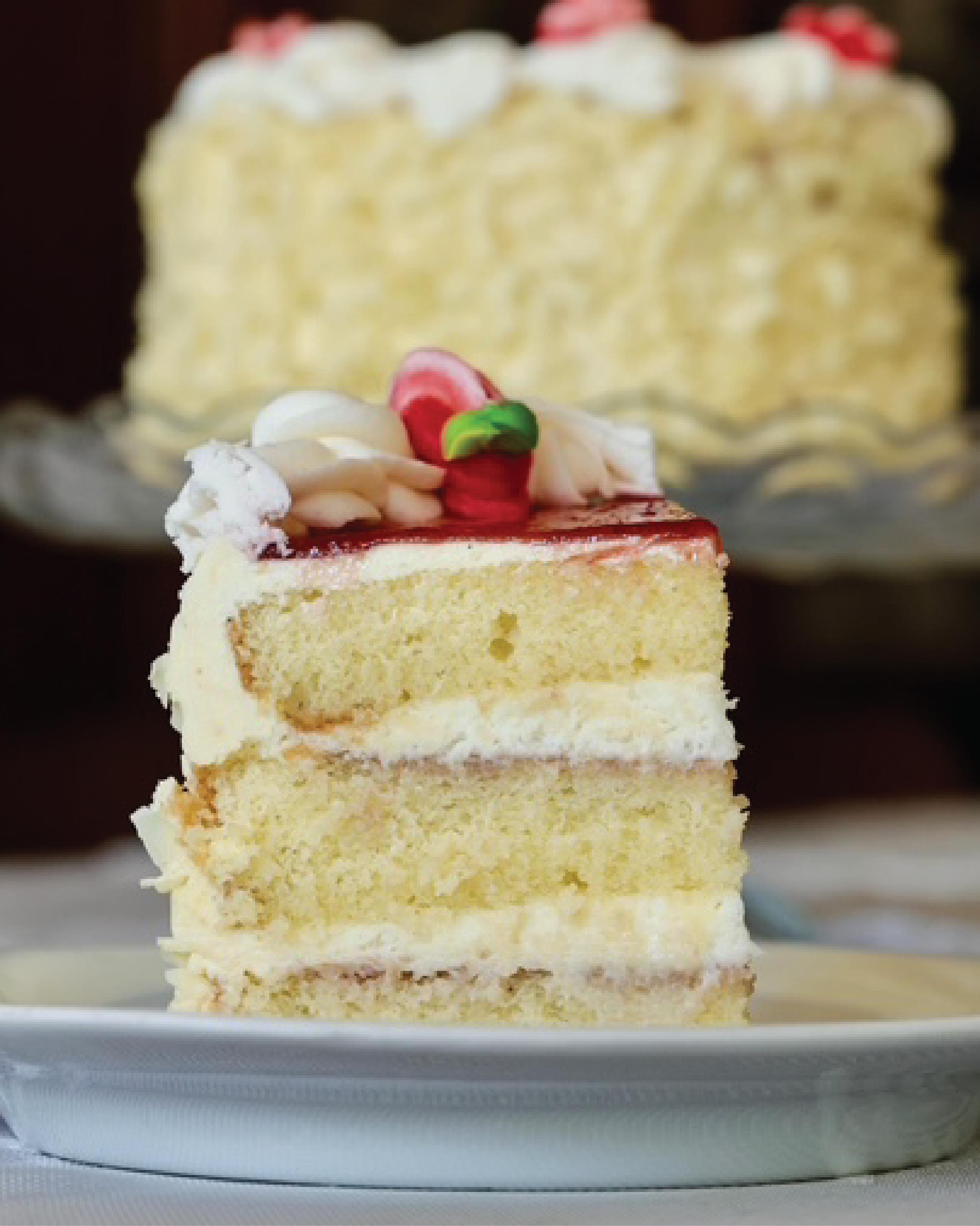 A slice of layered Bonbonerie Bumbleberry cake with white frosting and red fruit topping on a white plate, with a larger cake blurred in the background.