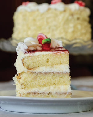 A slice of layered Bonbonerie Bumbleberry cake with white frosting and red fruit topping on a white plate, with a larger cake blurred in the background.