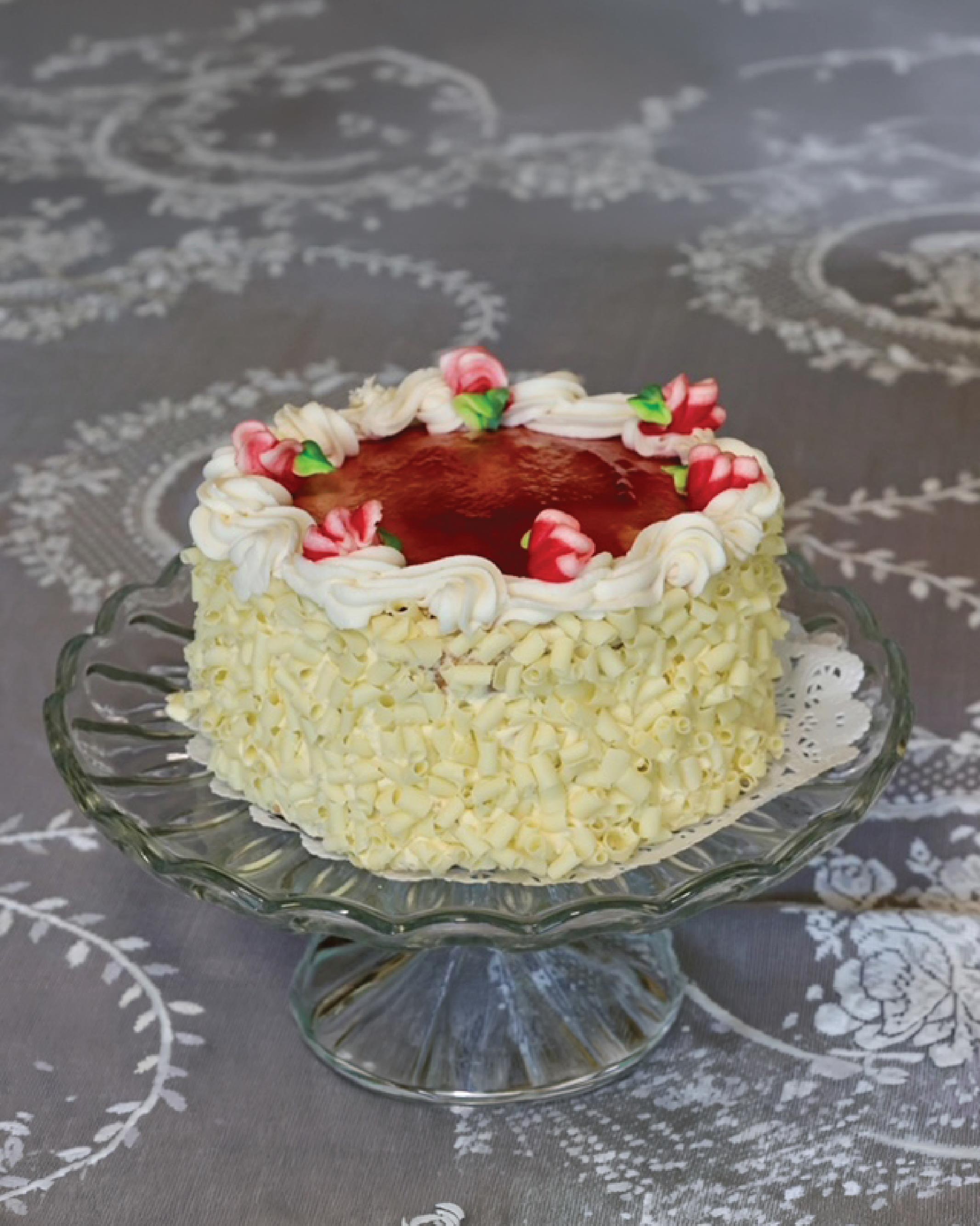 A round Bonbonerie Bumbleberry cake with white frosting, a glossy red top, and pink flower decorations, displayed on a clear glass pedestal plate over a gray patterned tablecloth.
