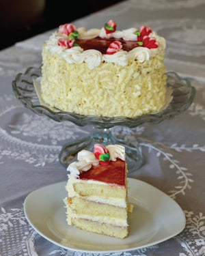 A white chocolate bumbleberry cake with red fruit topping is displayed on a glass pedestal stand, with a single slice placed on a white plate in front. The cake is decorated with piped white frosting and red-and-green candy accents, set on a patterned gray tablecloth.